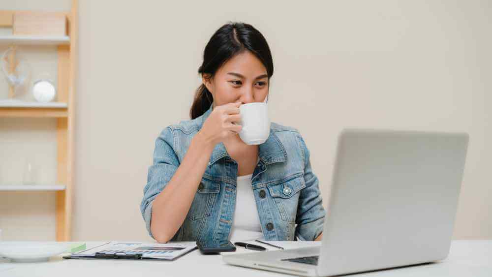 wanita asia sedang minum kopi sambil menghasilkan uang tambahan di depan laptop